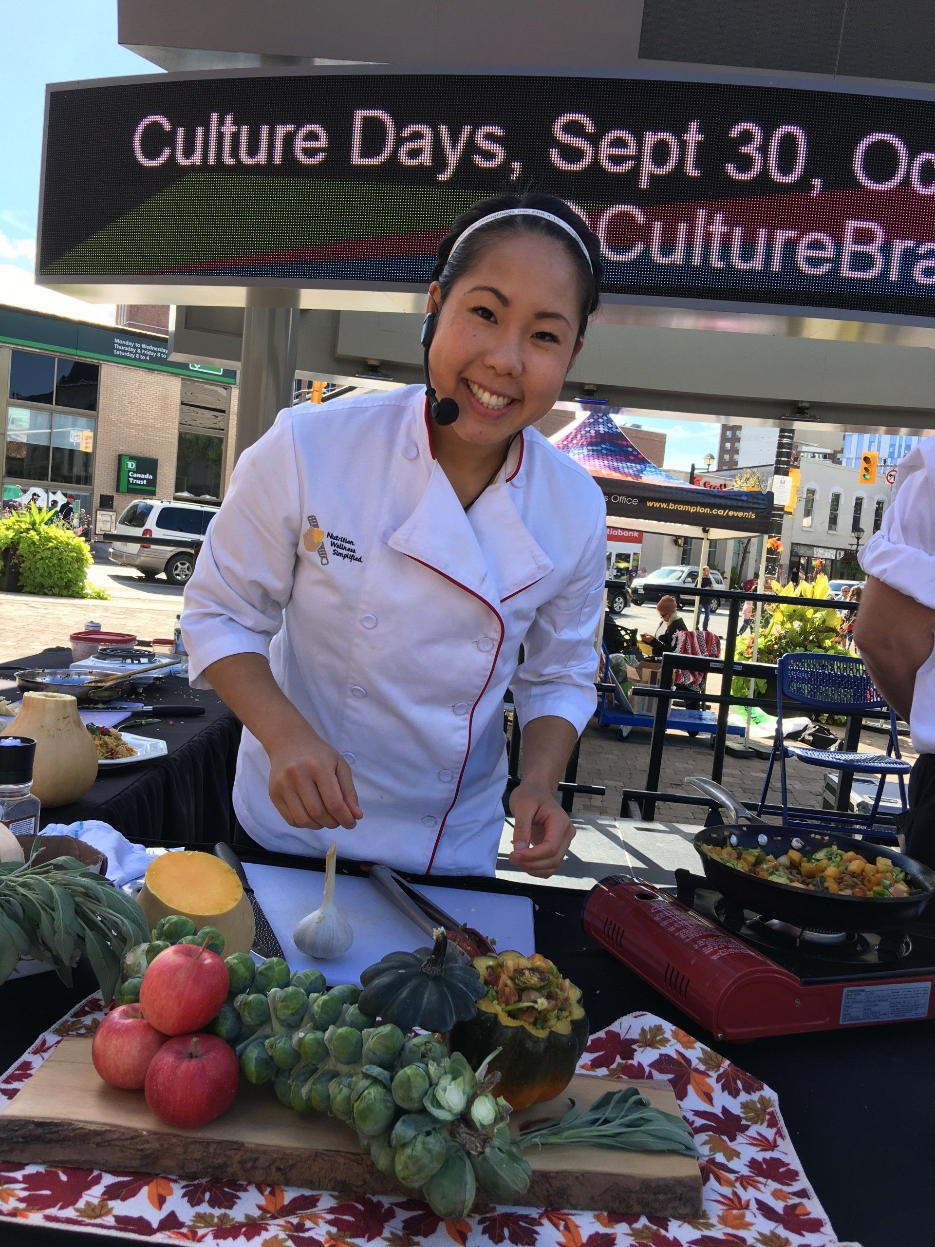 Chef preparing food at an outdoor event, smiling. Vegetables on display, Culture Days sign in the background.