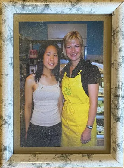Two women smiling, posing for a photo. One wears a yellow apron, the other a white top. Inside a shop.