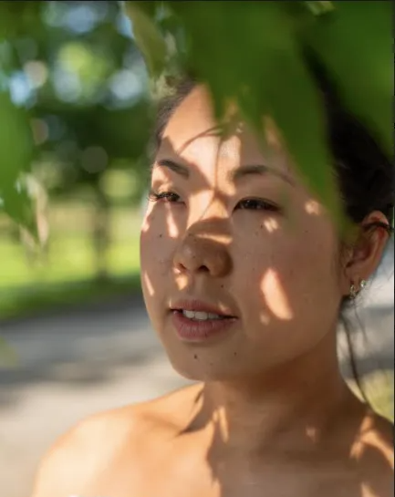 Woman's face in the sun, partially shaded by leaves. Sunlight dappled on her skin, soft smile.