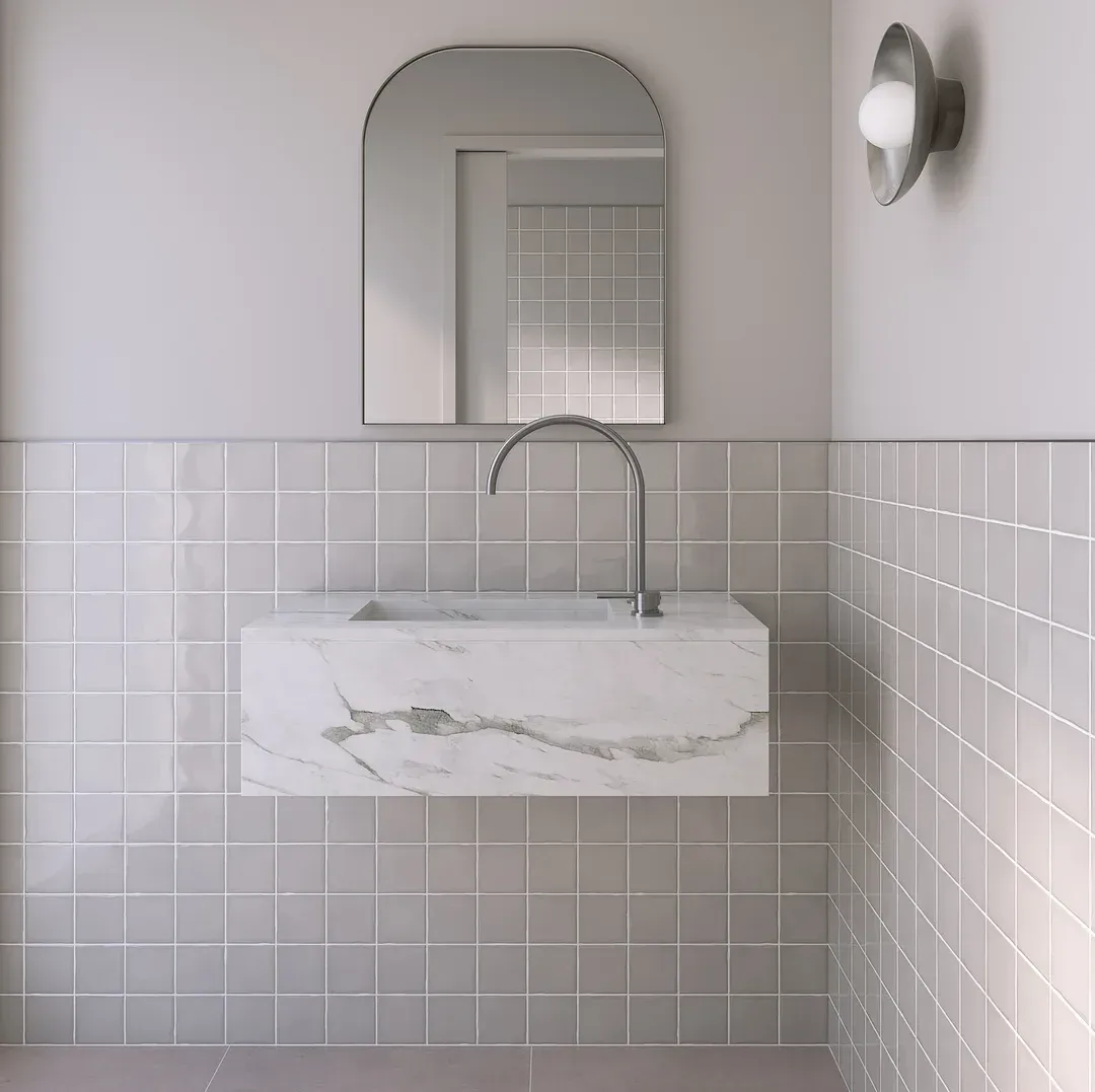 Modern bathroom with marble sink, arched mirror, gray tile wall, and sconce.