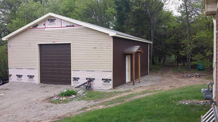 A large garage with a brown door is sitting in the middle of a grassy field next to a house.