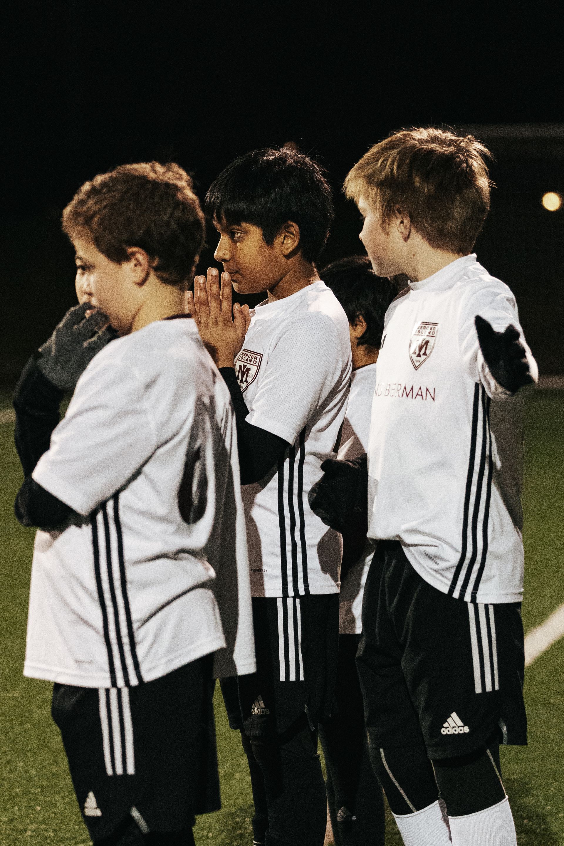 Three children in white soccer jerseys stand in a line on a field, one holding a microphone.