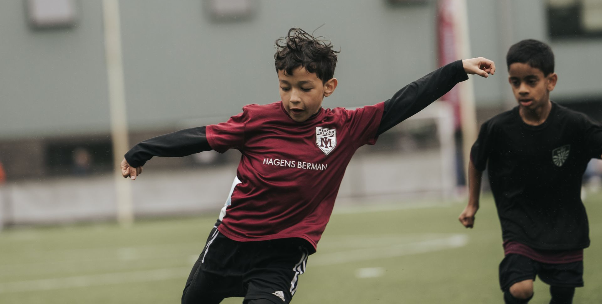 Young soccer player in maroon jersey dribbling a ball on a field, with another player in black nearby