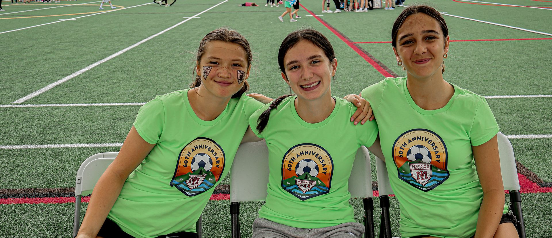 Three smiling teammates in neon green shirts sit on a field bench at a sports stadium.