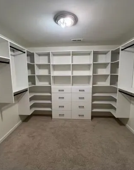 Empty white closet with built-in shelves and drawers, beige carpet, and a central light fixture.