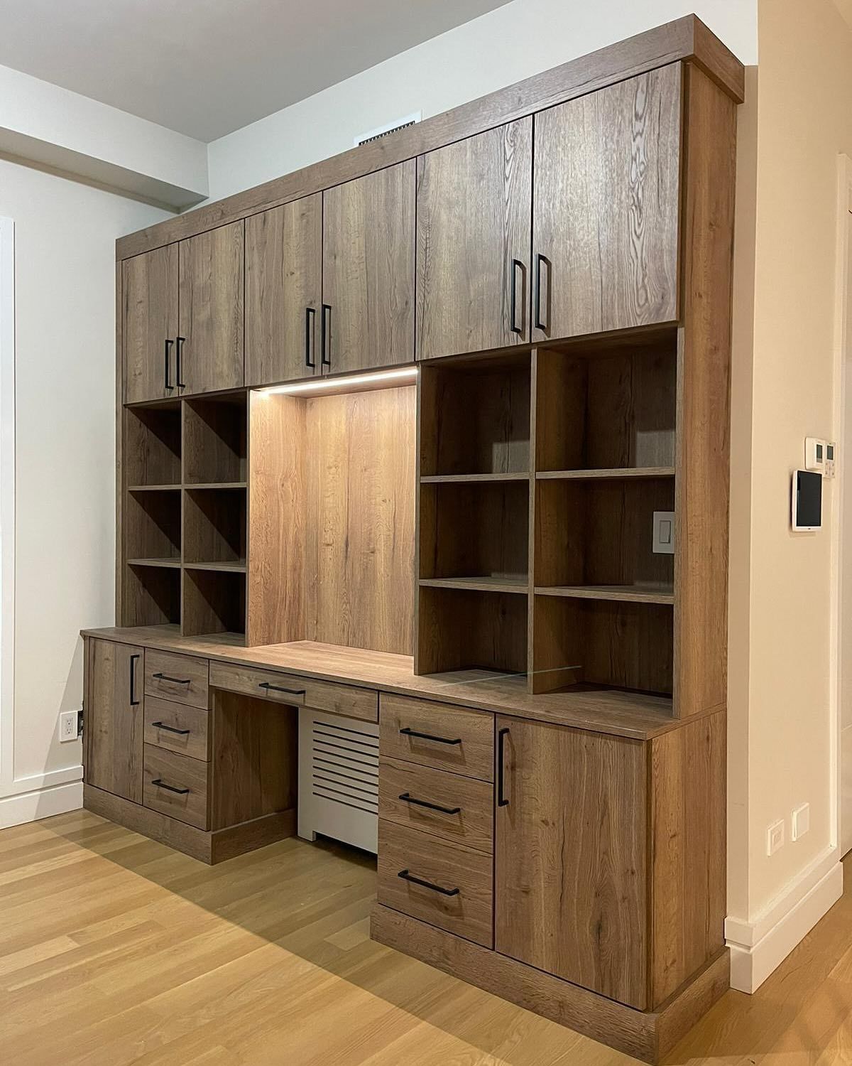 Built-in wooden desk and storage unit in a home office setting, with shelving, cabinets, and a desk area.