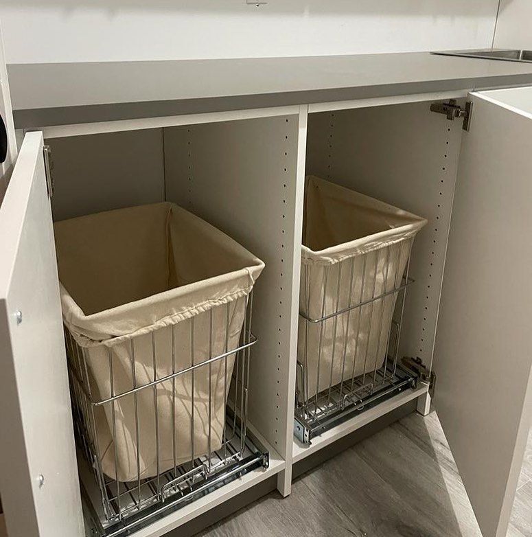 Laundry baskets in a cabinet, with pull-out wire frames and beige liners. White cabinet, open doors.