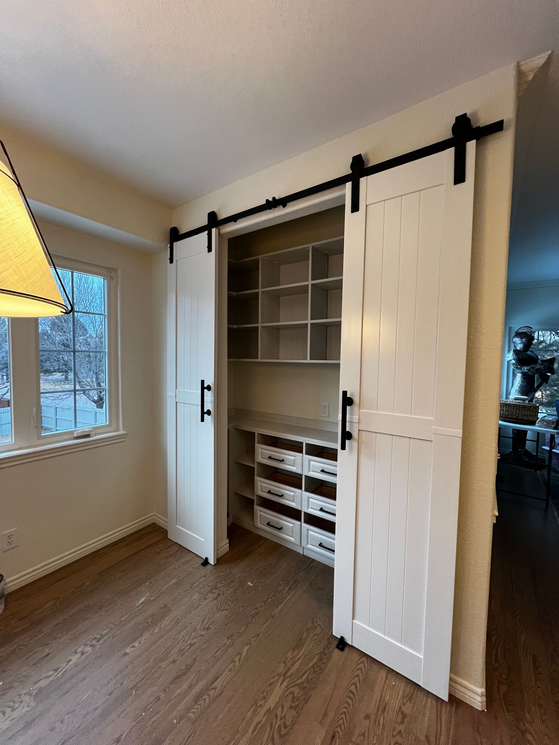 White pantry with barn doors open, revealing shelves and drawers. Black hardware, wood floor.