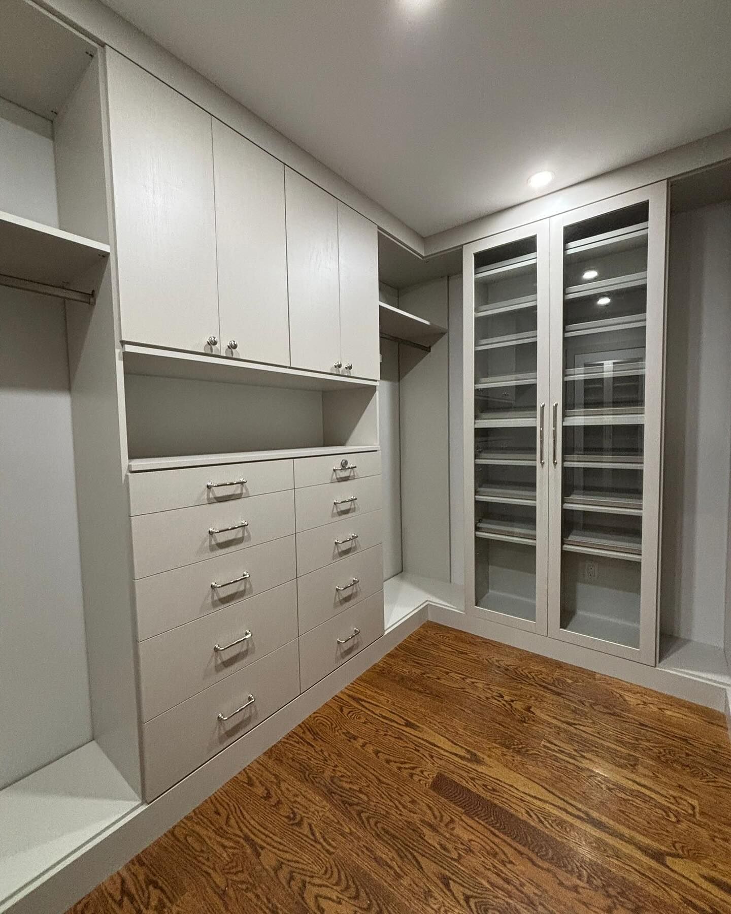 Walk-in closet with pale gray cabinetry, drawers, and glass-door shelving. Wood floor.