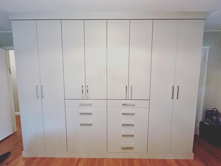 Built-in white wardrobe with multiple doors and drawers in a room with wood flooring.