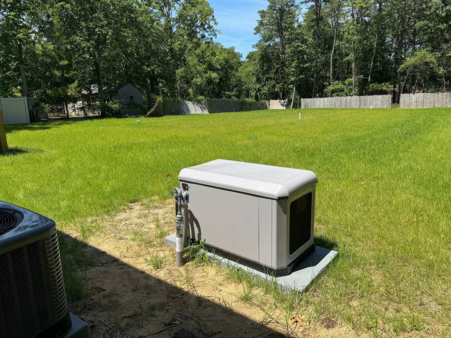 Gray generator box on a concrete pad in a grassy backyard with trees and a fence.