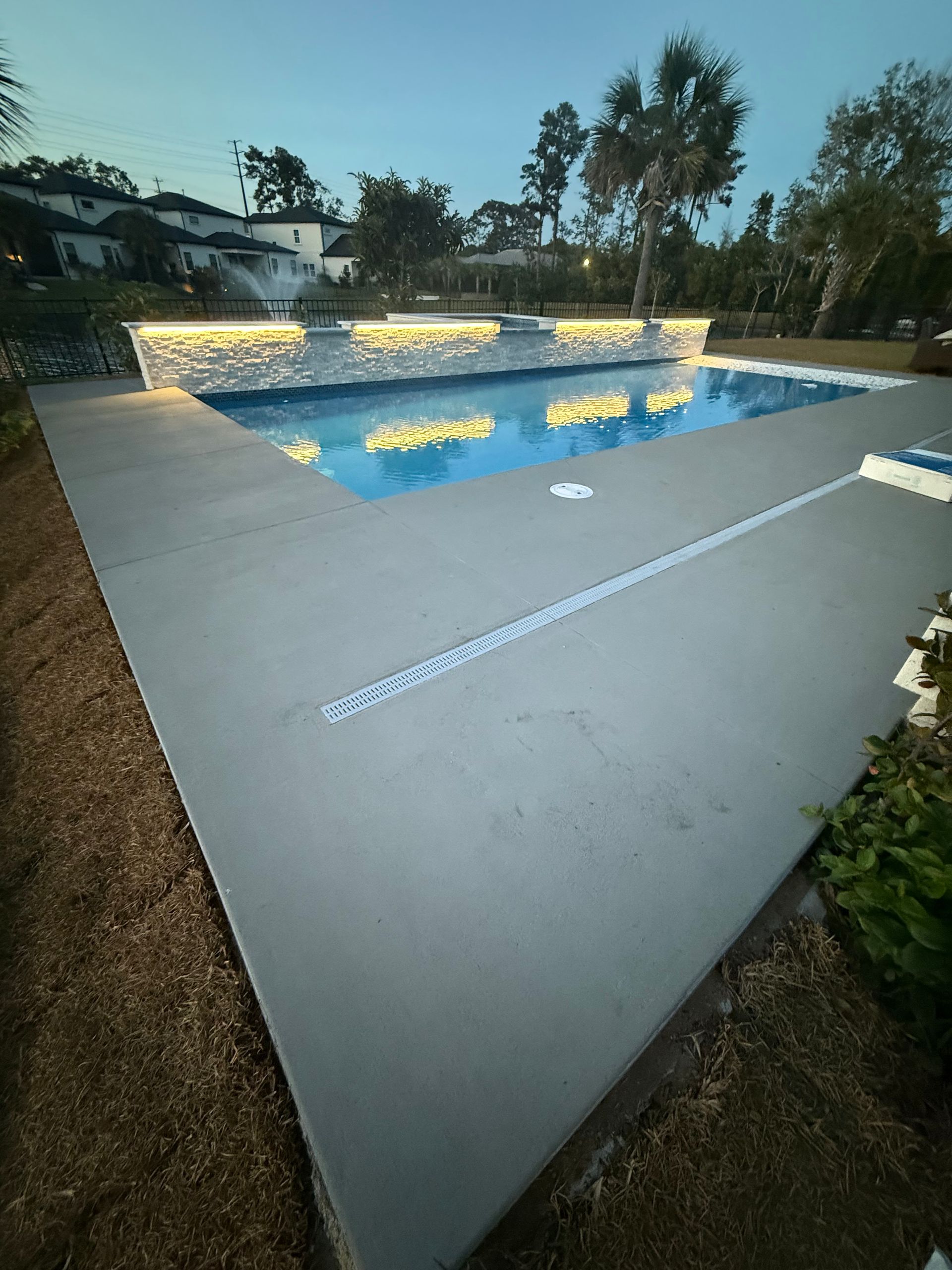 Lit swimming pool at night, surrounded by patio and outdoor seating area, with a house in the background.