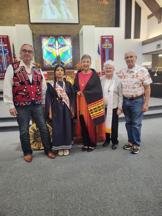 Five people in a church, some wearing Native American attire. Smiling, standing in front of a stained-glass window.