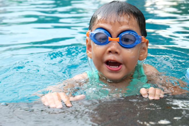 Child in blue goggles swimming in a pool, hands on the edge, with an open mouth.