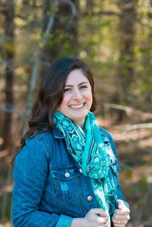 Woman in denim jacket, teal scarf, smiling outdoors in a wooded area.
