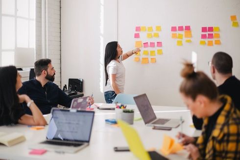 A team in a bright office reviews ideas on sticky notes; one woman points. Laptops are open on the table.