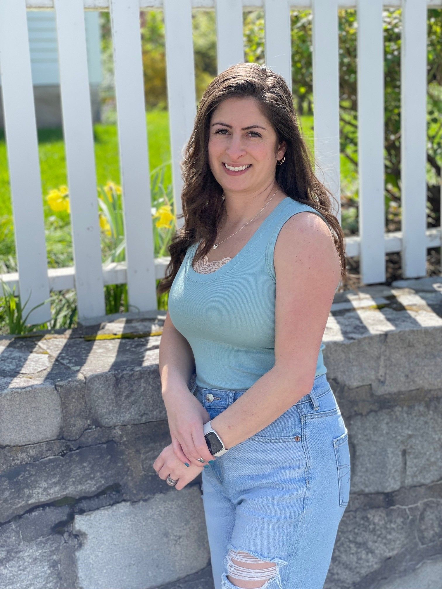 The owner Alexis in blue top and jeans, smiling, standing by a white fence and stone wall.