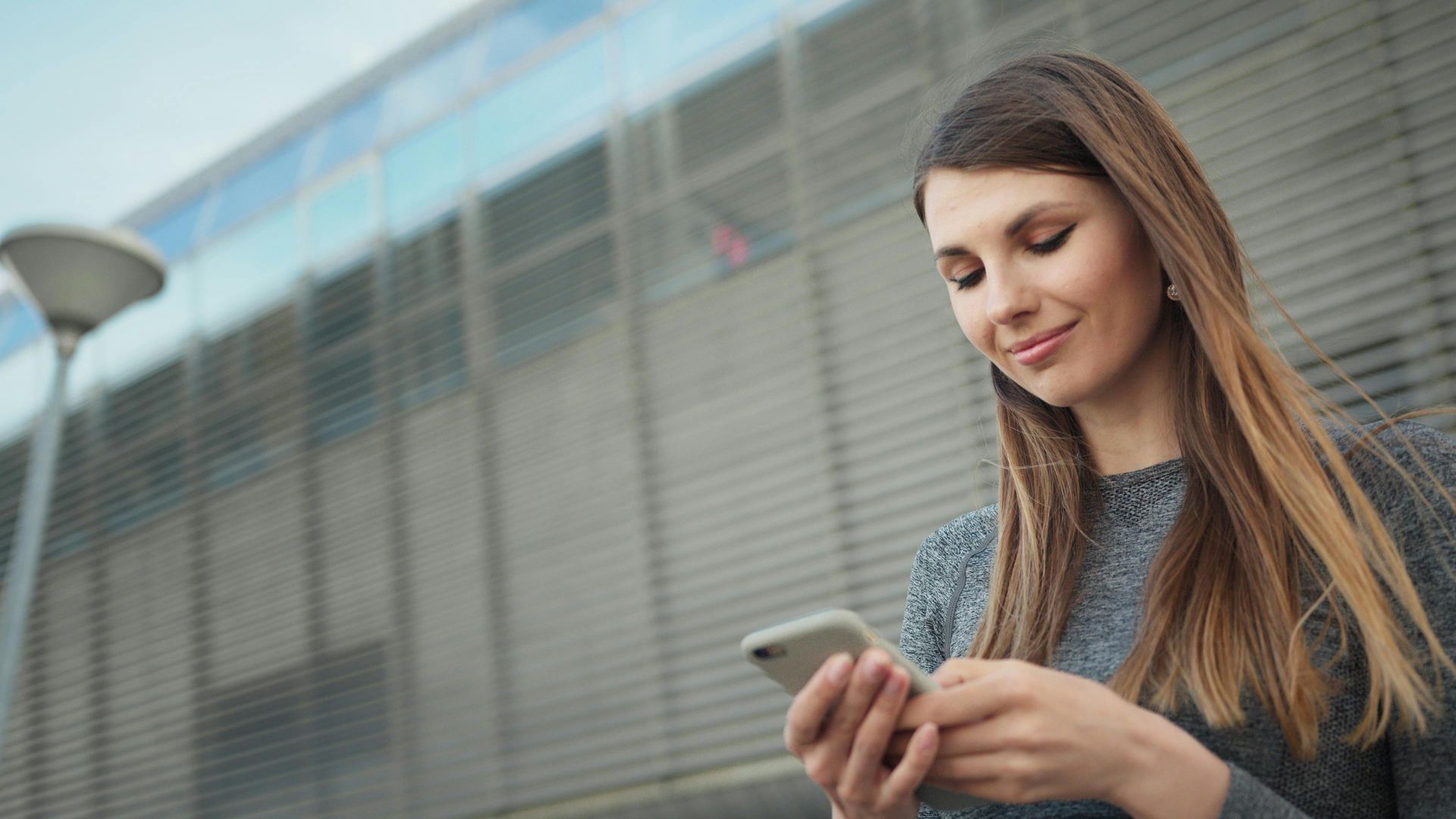 Woman smiling, looking at phone outdoors in front of a modern building.