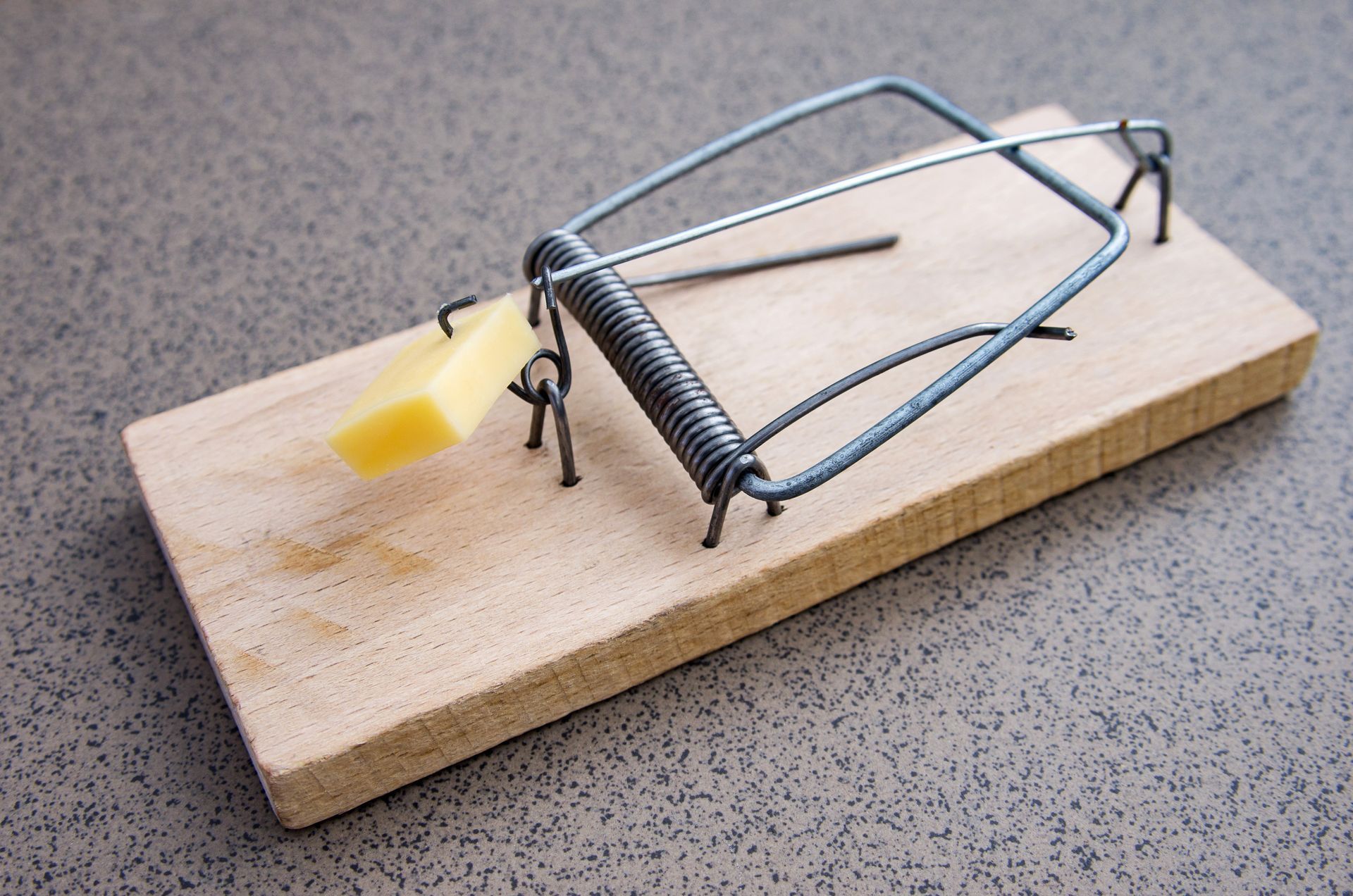 A metal spring-loaded mousetrap is placed on a wooden base set on a gray surface.