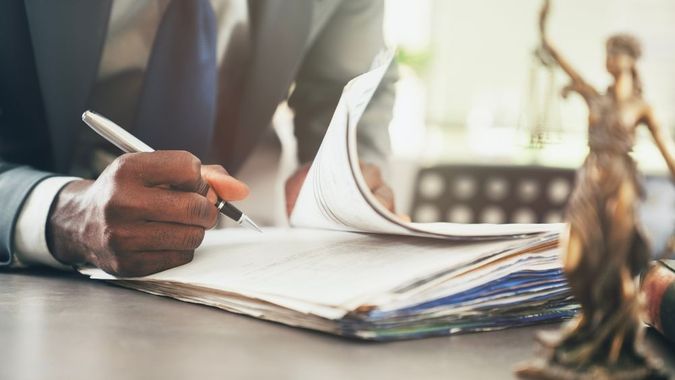 A person in a suit signing legal documents at a desk next to a statue of Lady Justice.