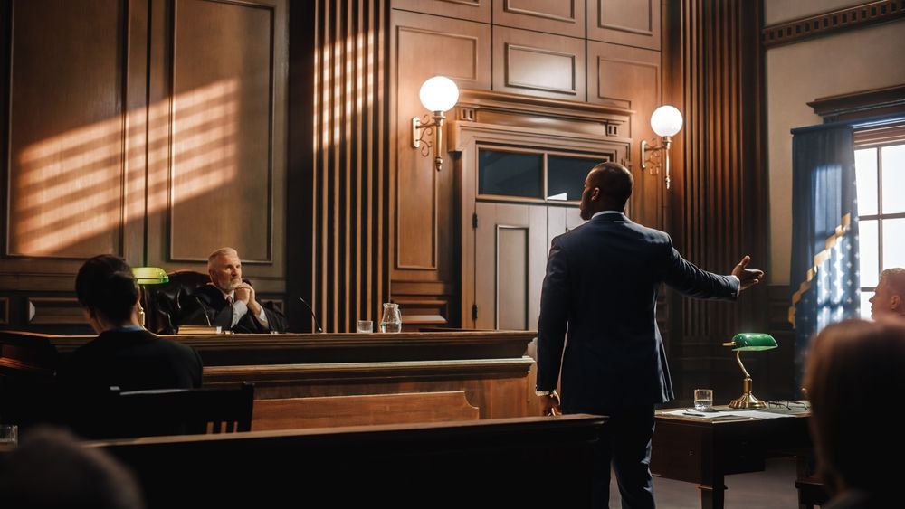 A lawyer stands and gestures in a courtroom while a judge sits at the bench, bathed in soft light from nearby windows.