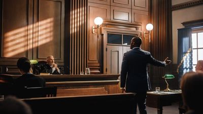 A lawyer stands and gestures in a courtroom while a judge sits at the bench, bathed in soft light from nearby windows.