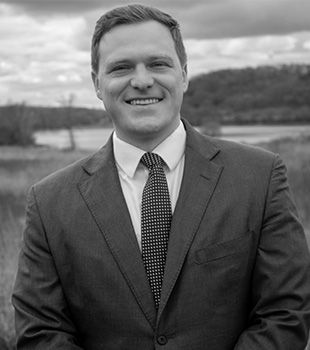 Headshot of a smiling person in a suit and tie, standing outdoors in front of a rural landscape.