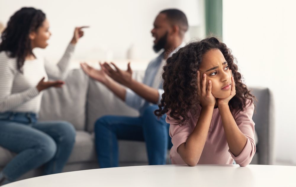 A child sits at a table looking sad while their parents argue in the blurred background.