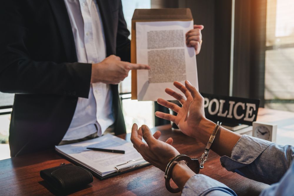 A person in a suit points to a document while a person in handcuffs sits opposite them at a desk marked with a police sign.