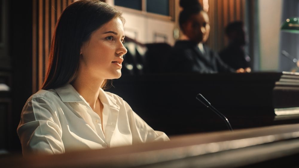 A woman in a white collared shirt speaks into a microphone while seated at a wooden desk in a courtroom setting.