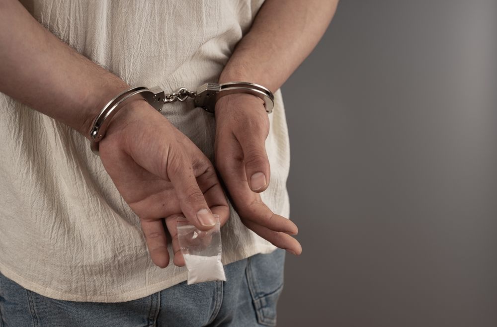 A person in handcuffs stands with their hands behind their back, holding a small plastic bag filled with white powder.