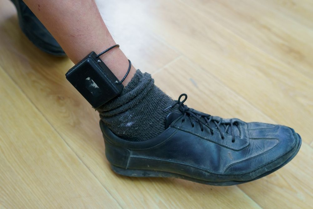 A person wearing a black ankle monitor device over a dark sock and shoe on a wooden floor.