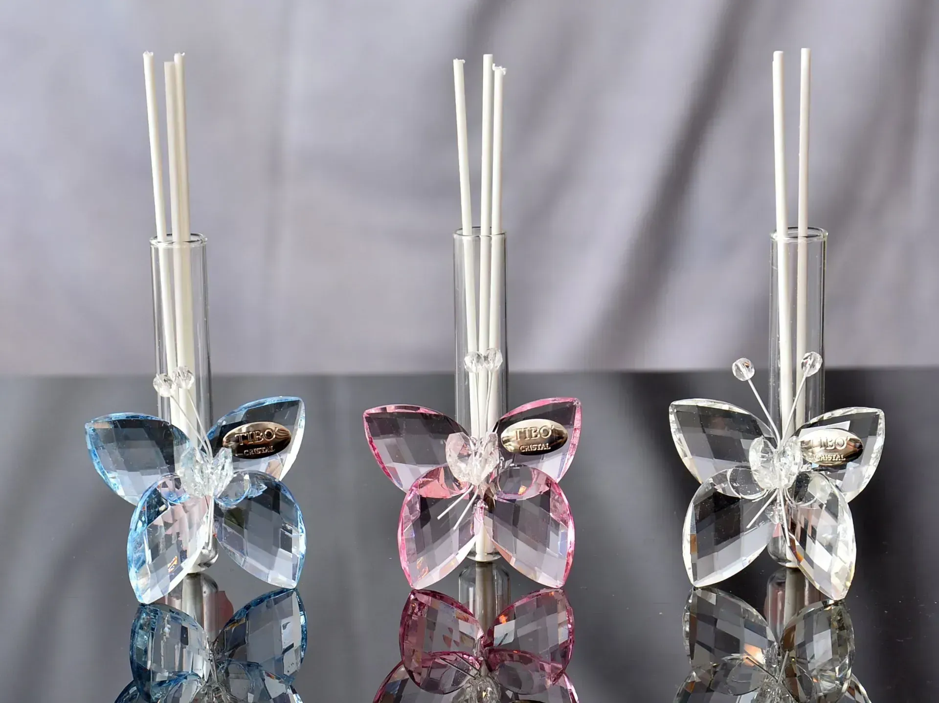 Tre ornamenti a forma di farfalla in cristallo con tubi di vetro che contengono canne, nei colori blu, rosa e trasparente.