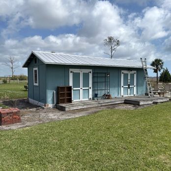 Blue rectangular house with white doors, metal roof, and wooden deck under a cloudy sky.