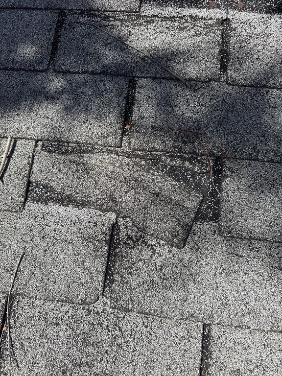 Close-up of weathered asphalt roof shingles. One shingle is partially detached, with dark shadows.