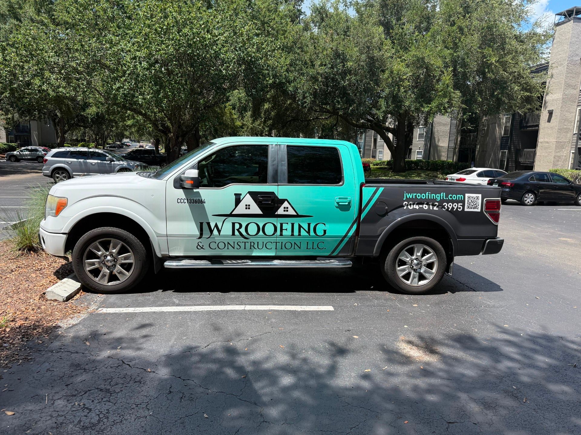 White and teal JW Roofing truck parked in a lot, logo on side, with trees and cars in background.