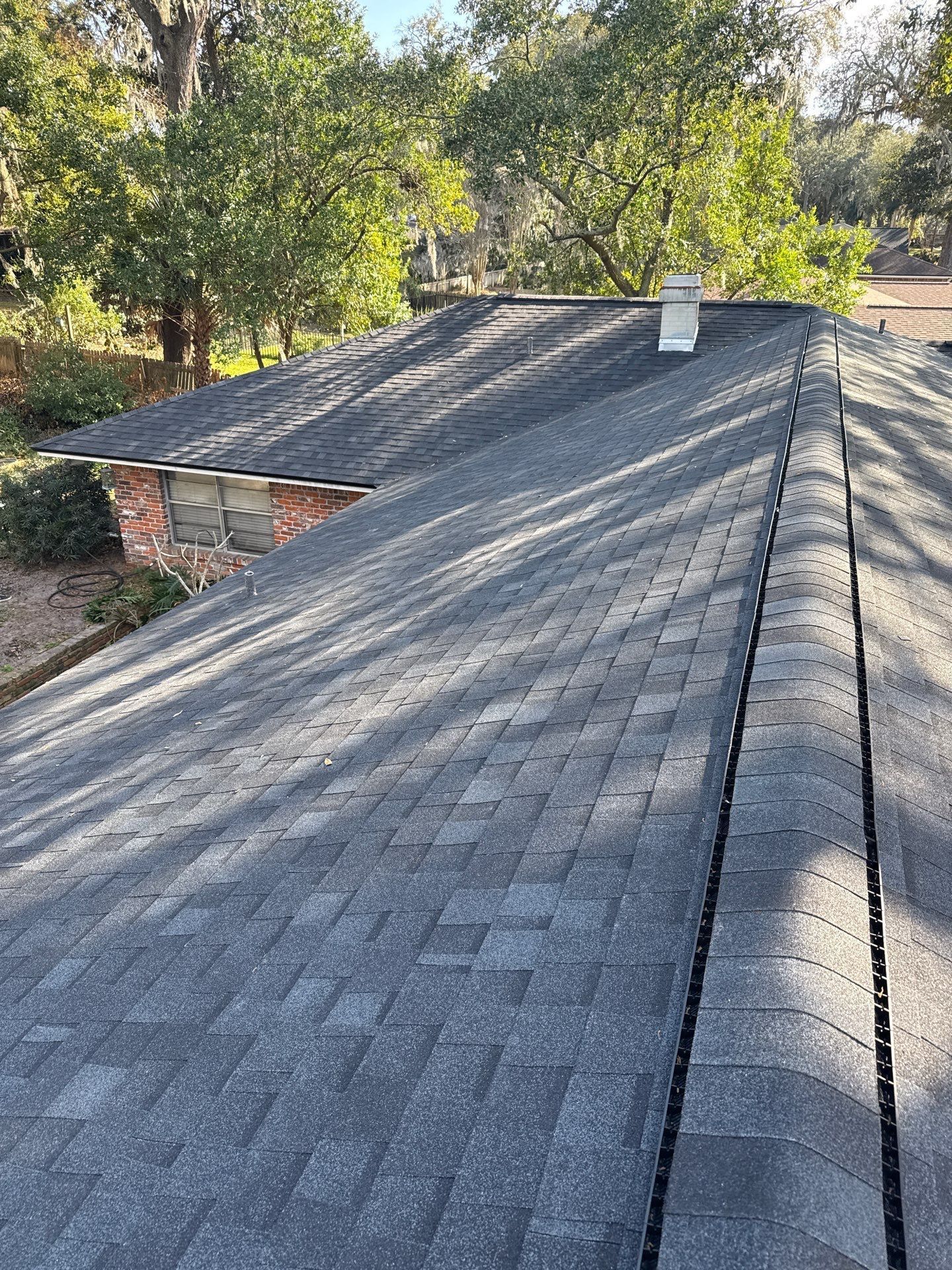 Dark gray asphalt shingle roof on a house with a chimney, trees in the background.