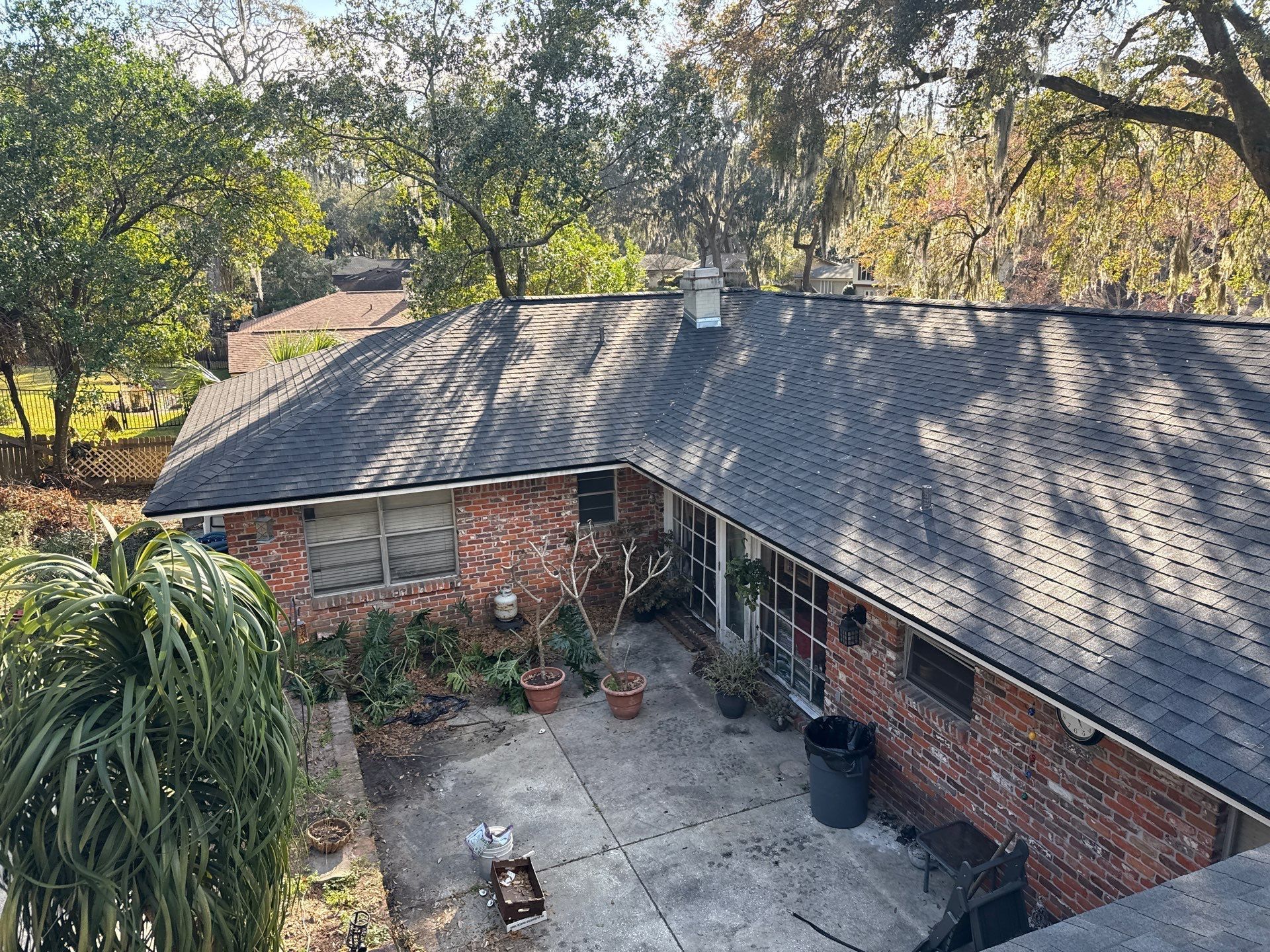 Overhead view of a brick house with a dark shingled roof, a patio, and trees in the background.