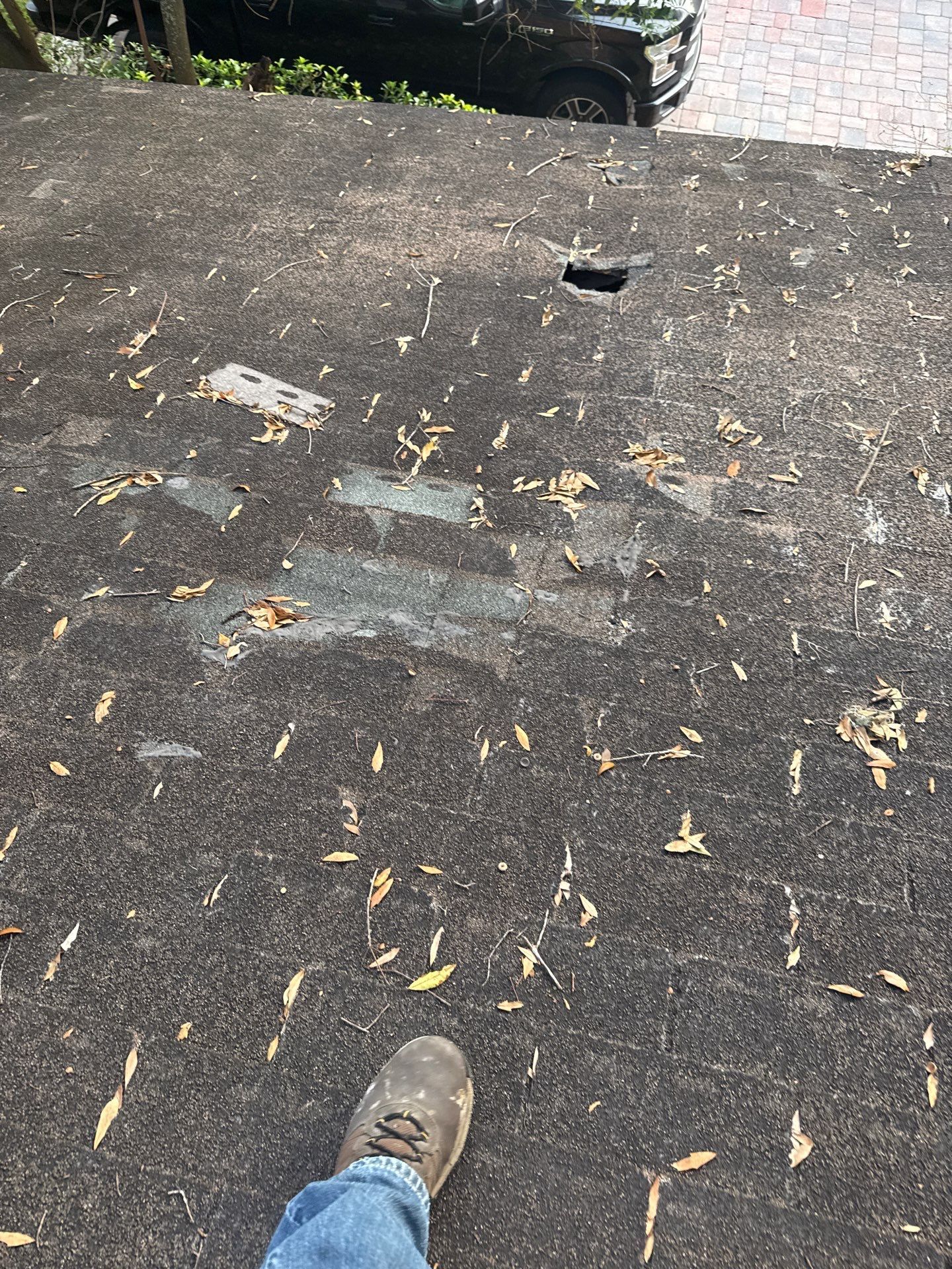 Foot on a dark, textured surface with leaves and a drain. A car and red brick are visible in the background.