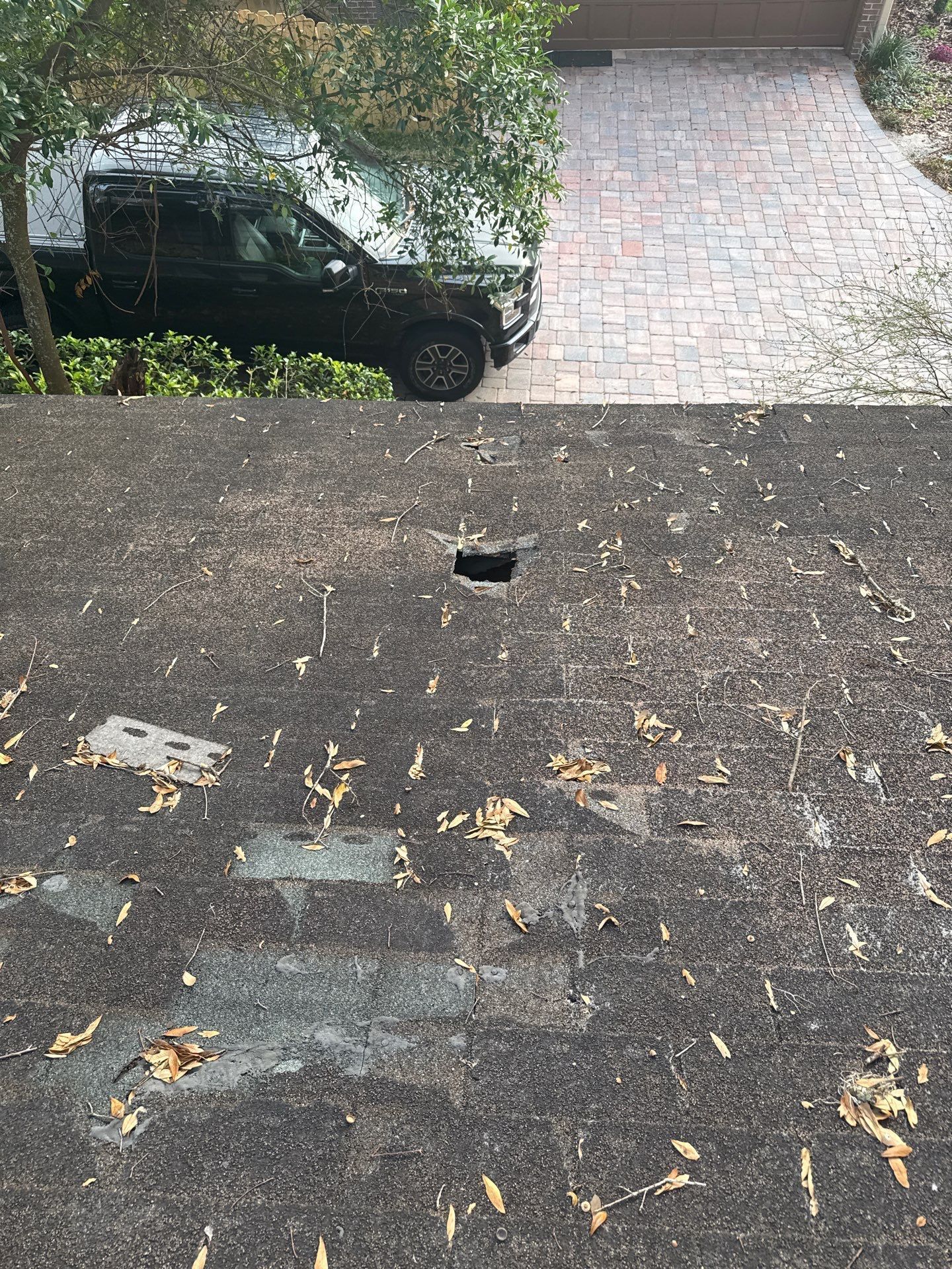 Dark asphalt with a small hole. A black car is parked nearby on a brick driveway, with a tree in the background.