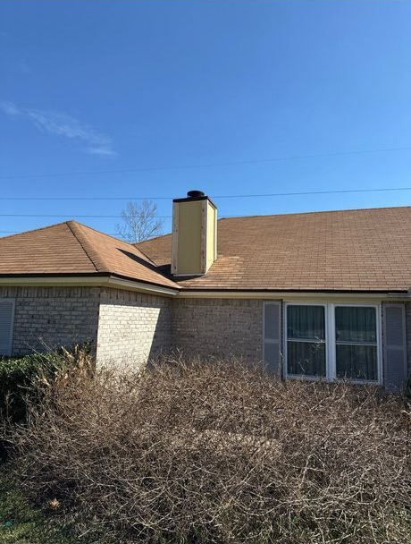 A beige brick house with a brown roof and chimney, overgrown bushes in front, on a bright blue day.