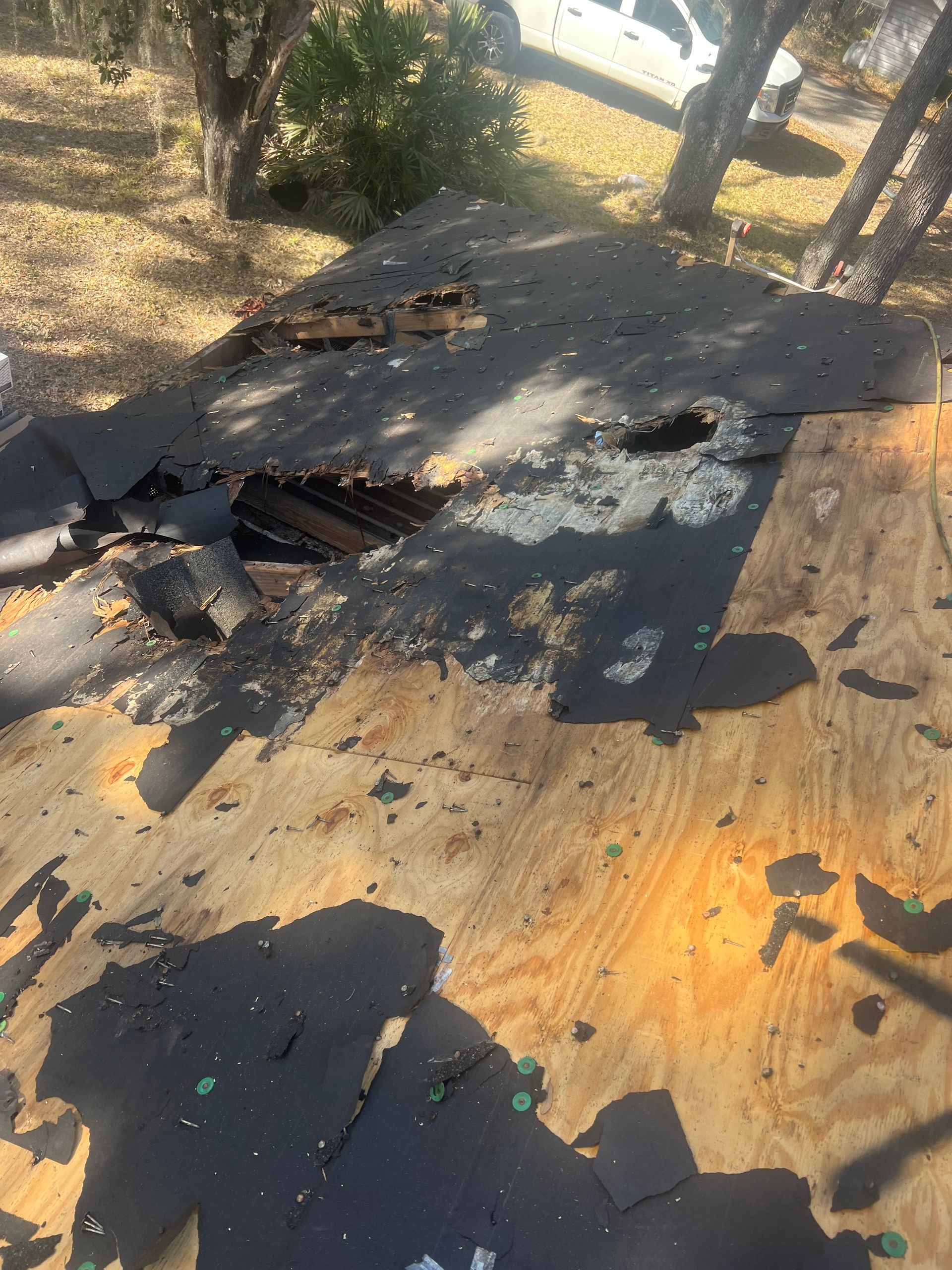 Damaged roof exposing plywood and a hole. Debris scattered, with trees and a vehicle in the background.