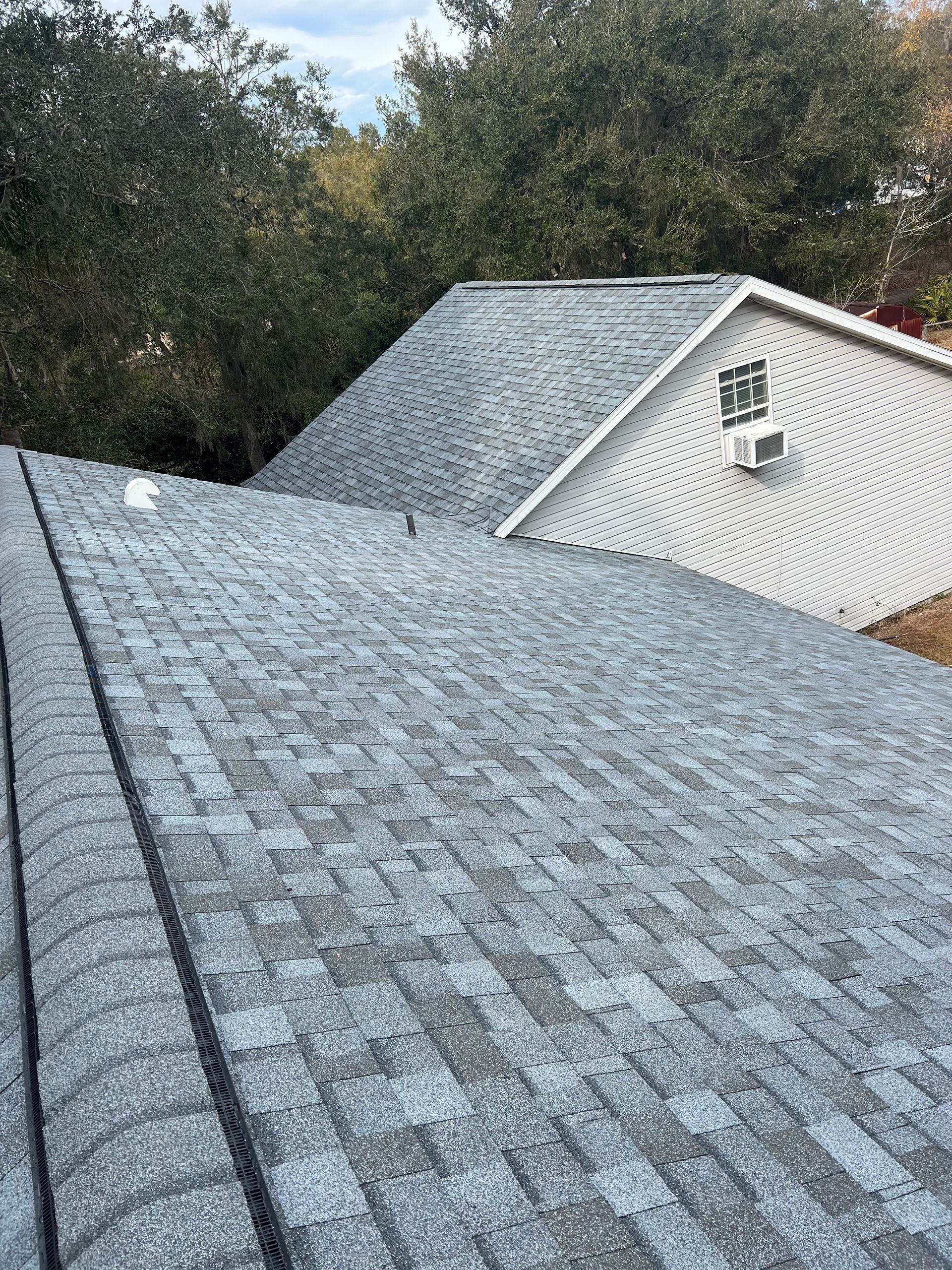 Gray shingle roof of a house with trees in the background and a white gable.