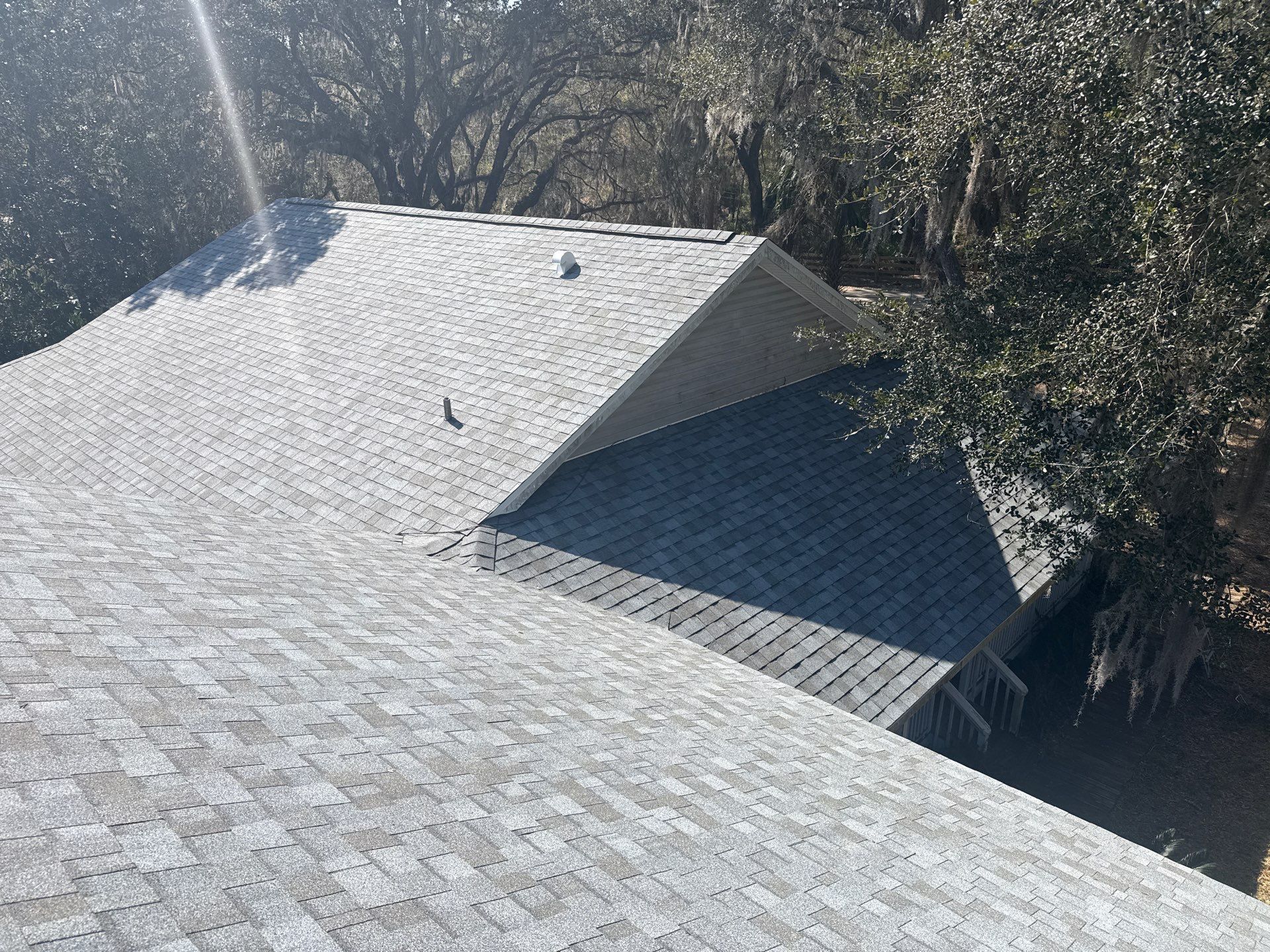 Gray shingle roof of a house, surrounded by trees, under a bright sun.