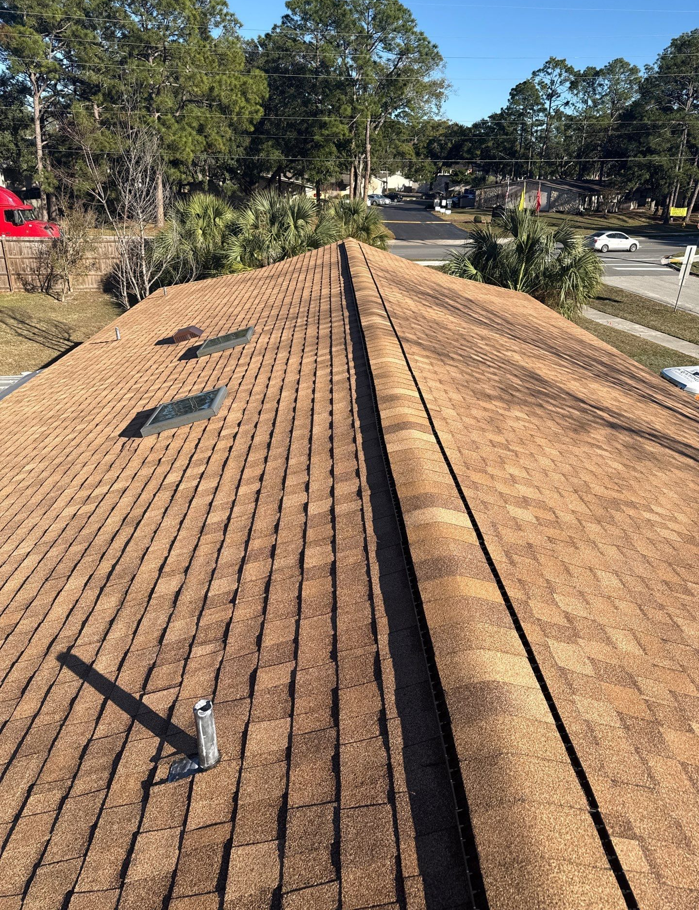 Brown shingle roof with two skylights and a vent pipe, overlooking a road and trees on a sunny day.