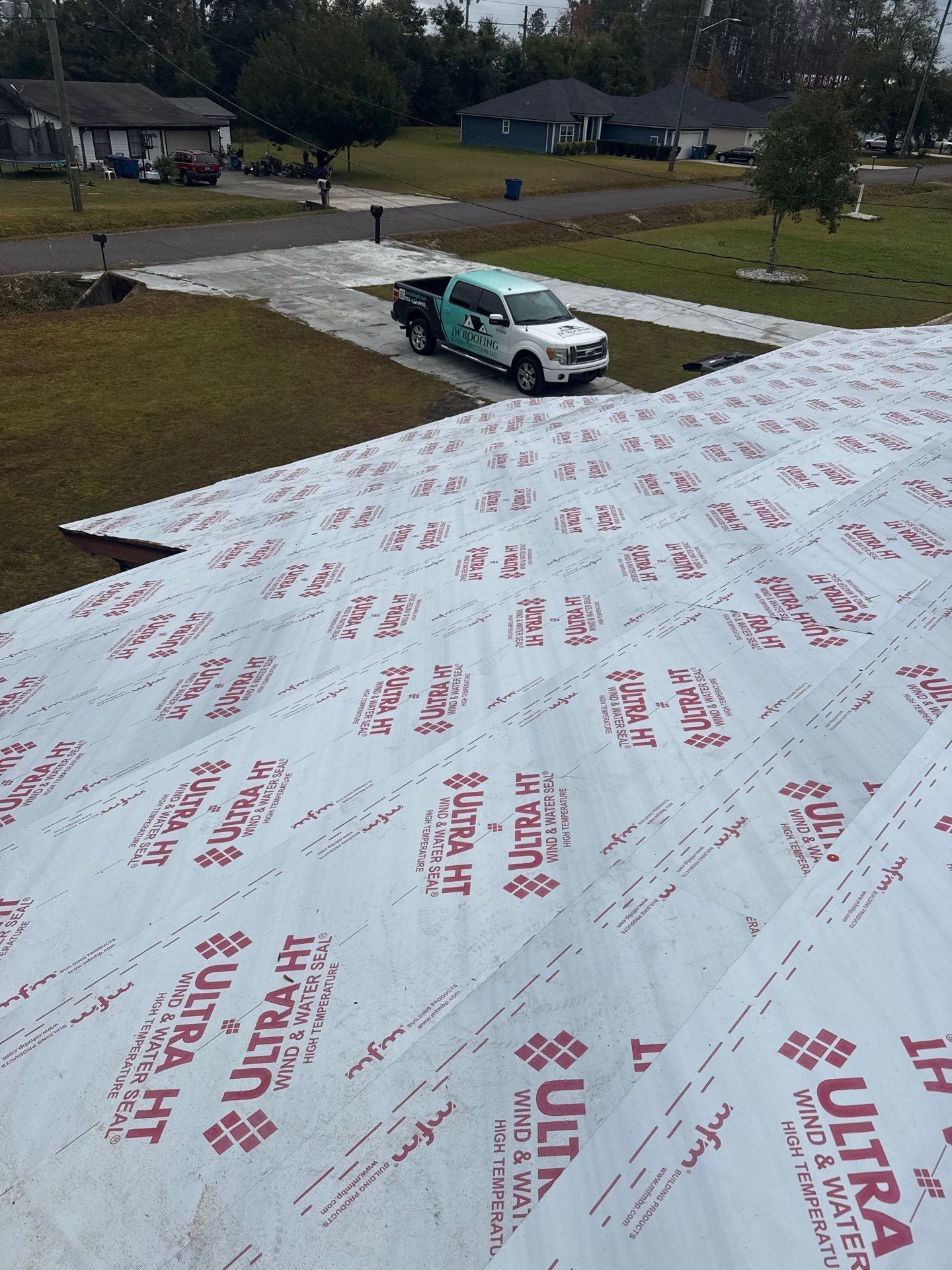 White roofing underlayment on a roof with a work truck parked below, and houses in the background.