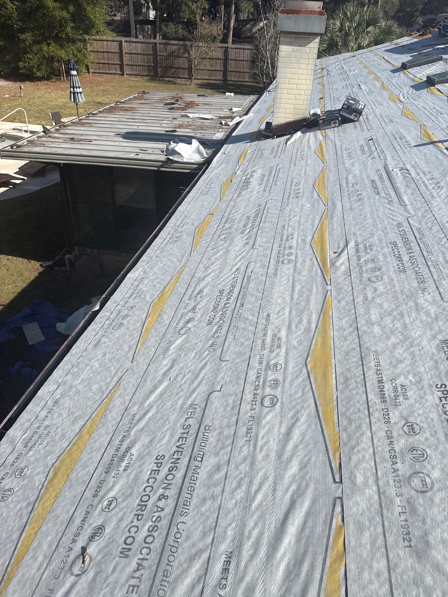 Rooftop partially covered with grey underlayment material, near a brick chimney, with an older roof visible.