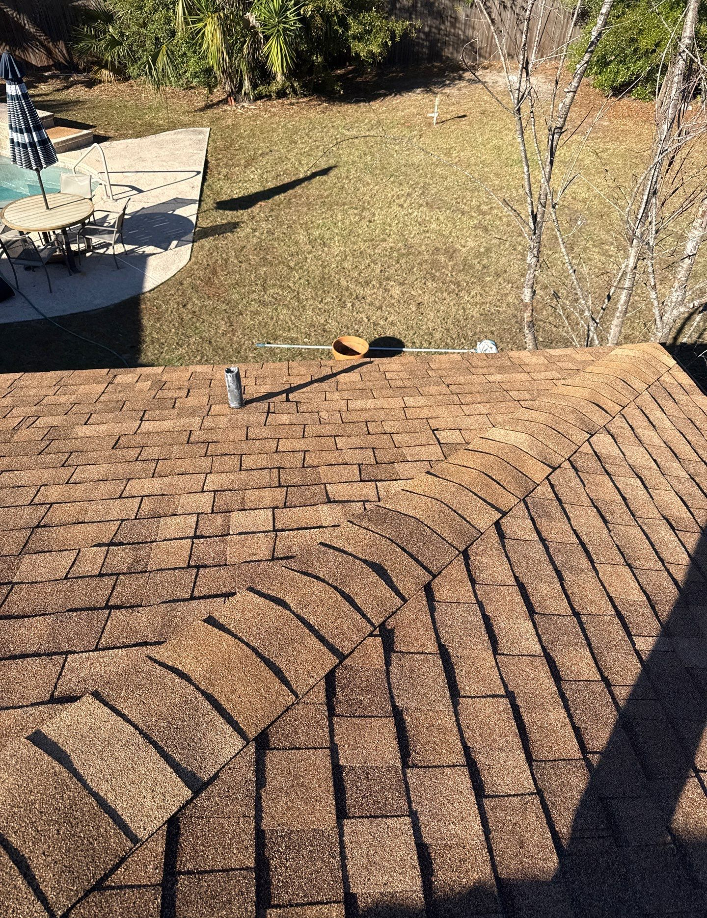 Brown asphalt shingle roof with a curved edge; a metal pipe runs across the peak. Backyard with grass and pool in background.