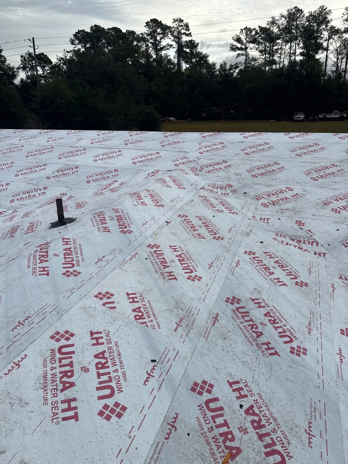 Roof with Ultra HT underlayment installed, flat surface, dark trees and cloudy sky in the background.