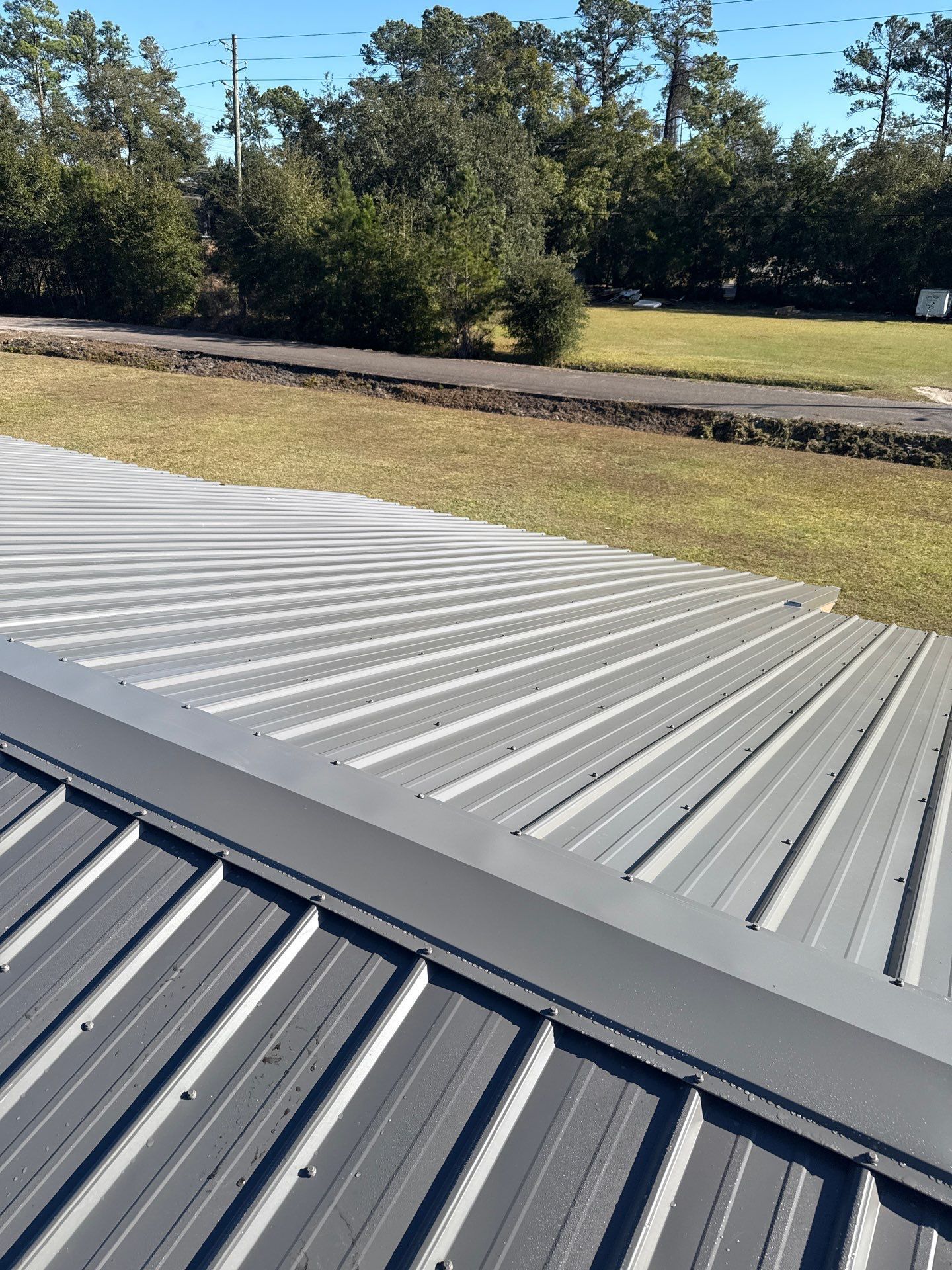 Close-up view of a gray metal roof with ribbed panels, contrasting with a grassy field and trees in the background.